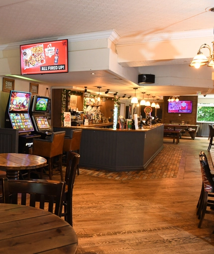 Interior dining area of a pub with a bar, a dartboard, a few TVs and gambling machines.