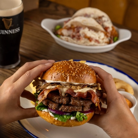 Two hands holding a Big Cheese Melt Burger over a plate on a wooden table.