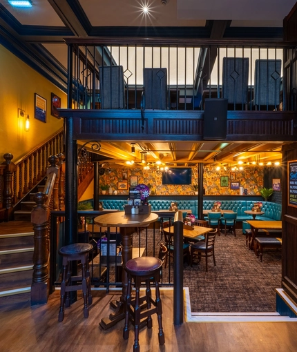 A view of a ground floor interior restaurant seating area at the Old School House, a ground floor games area with gaming machines, and a wooden staircase leading up the upper floor and balcony above.