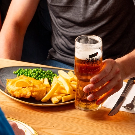 A hand holding a pint of Carling next to a plated portion of fish and chips, at an indoor table.