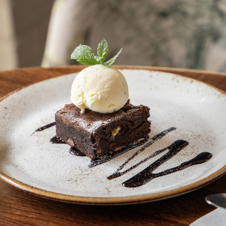 A triple chocolate brownie served on a plate and topped with a scoop of ice cream and chocolate drizzle. The plate sits on a wooden table next to cutlery on a napkin.