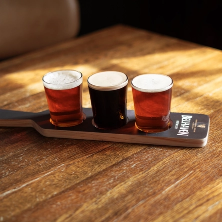 Three half pint glasses of ale or craft beers sitting in a paddle shaped drinks holder on a wooden table inside a pub.