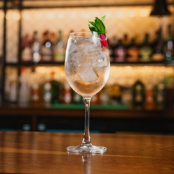 A close up view of a non-alcoholic Hugo Spritz cocktail served in a wine glass with ice, mint, and a lime wedge. The glass stands on the bar inside a pub.