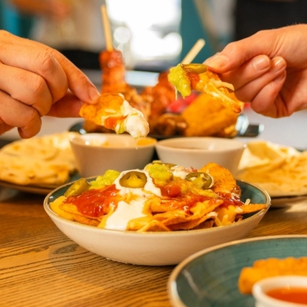 Two hands reach into a shared plate of loaded nachos topped with melted cheese, jalapeños, guacamole, salsa and sour cream. The table also features dipping sauces, flatbreads, crispy snacks and other small plates, creating a casual sharing-dishes dining scene.