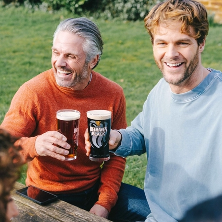 Two people sitting at a wooden table in a sunny beer garden, raising their glasses of Belhaven Best and Belhaven Black.