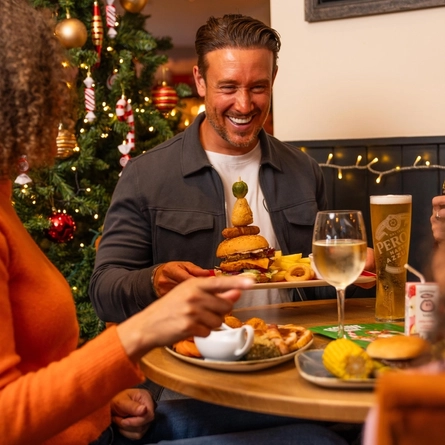 A festive dining table in a Christmas-themed restaurant, with a decorated Christmas tree featuring red and gold baubles, candy cane ornaments and warm fairy lights in the background. One diner holds a plate topped with a stacked burger, onion rings and chips, while other diners sit around the table with food and drinks. White wine, beer, fruit juice and Christmas-themed table decorations are arranged on the table, creating a warm seasonal dining setting.