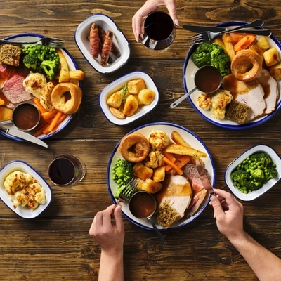 An image showing the torso of a person sat at an indoor table. Their hands are holding a knife and fork over a plated roast dinner.  There are several plated roast dinners on the table.