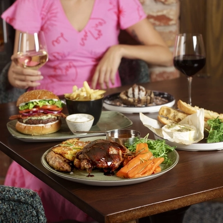 A person in a pink dress holds a glass of white wine at a table with assorted dishes including a burger, grilled meat, bread with cheese, and dessert.