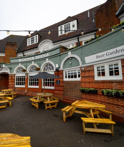 The exterior beer garden seating area at the Wooden Bridge in Guildford, with wooden picnic tables, shade umbrellas, and string lights overhead.