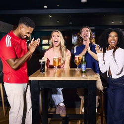 An image showing 4 friends at a table enjoying drinks and celebrating while watching on a football match within the interior seating area at a Flaming Grill venue.