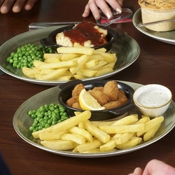 A round table filled with main dishes, including scampi and chips and a pie with mash and peas. Several drinks are placed around the table. The hands and arms of multiple people seated together are visible as they reach toward their food and raise their drinks.