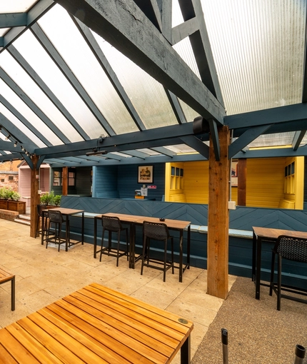The paved, exterior seating area underneath a large wooden pergola at The Talbot; with a TV on the wall, and string lights hanging above the tables and chairs.