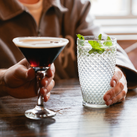 Two people holding cocktails which sit on a wooden table. One cocktail is served in a martini glass and the other is served in a highball glass and garnished with mint leaves.