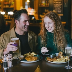An image of a 2 friends enjoy Fish & Chip dishes with drinks sat within the interior restaurant seating area at an Urban Core Venue.
