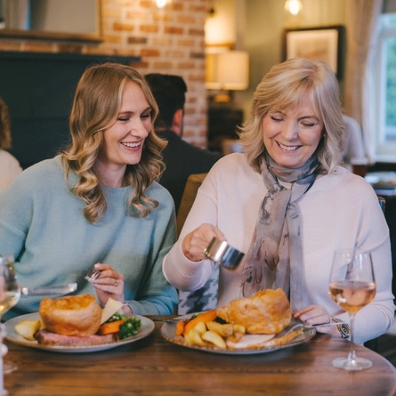 A lifestyle image of 2 ladies enjoying Mothers Day roast main dishes and drinks within the interior restaurant seating area at a Pub & Grill venue.