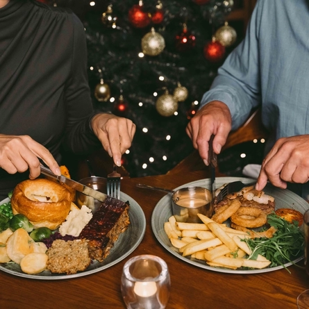 A plate of Christmas dinner and a plate of steak with prawns, fries, onion rings, tomato, salad, and a pot of sauce; sitting on a wooden restaurant table along with a tealight and glasses of white wine and mulled wine. Two people sitting at the table are cutting up the food with their knives and forks; and a Christmas tree is visible in the background.
