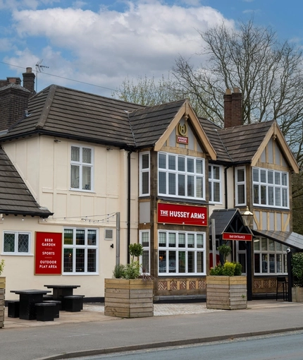The exterior facade, signage, and seating area of The Hussey Arms in Brownhills, with picnic tables, planter boxes, and string lights.