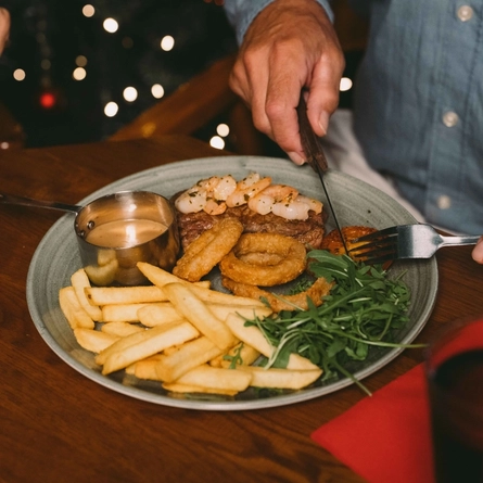 A close up view of steak with prawns, fries, onion rings, tomato, salad, and a pot of sauce; served on a plate which sits on a wooden restaurant table. Another plate of food, a tealight, a serviette, and a glass of mulled wine also sit on the table; and two people sitting at the table are cutting up the food with their knives and forks.