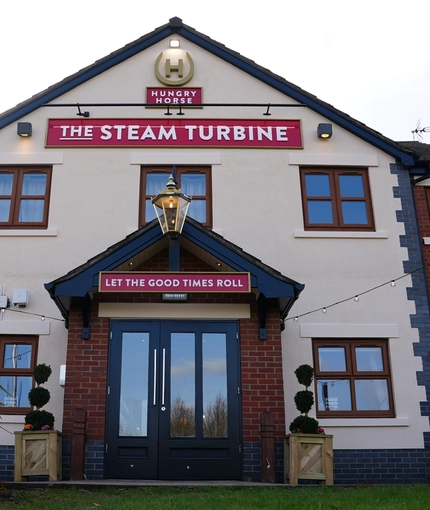 The exterior facade and signage of The Steam Turbine, with potted topiary trees on either side of the doorway, and string lights overhead.