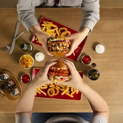 The hands of two people seated at a wooden table each holding a burger.  On teh table in front of each person are plates of fries and onion rings.