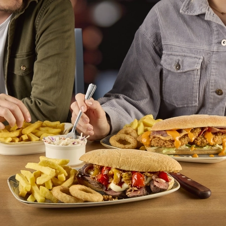 A steak sub sandwich and a chicken sub sandwich, each served on a plate with chips and onion rings. The plates sit on a wooden table along with a ramekin of coleslaw and a small roasting dish of chips which two people sitting at the table are eating from. 