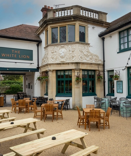 The exterior facade, signage, and beer garden seating area of The White Lion, with wooden picnic tables and string lights overhead. 