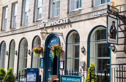 The exterior facade, signage and seating area of The Advocate in Edinburgh, with hanging flower baskets on either side of the entrance.