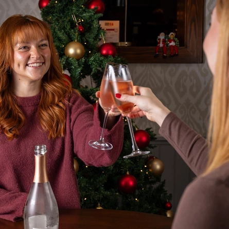 Two women stood at an indoor table,  holding glasses of sparkling wine.  A decorated Christmas tree is behind them.