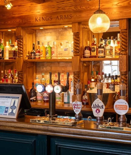 A close up view of the wood panelled bar inside The Kings Arms in Sandford-On-Thames, with spherical lights hanging above the counter, and bottles of alcoholic spirits on wooden shelves behind the bar.