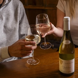 Two people sitting at a wooden table inside a pub, clinking together their glasses of wine. A bottle of Louis Robin Chablis wine stands on the table.