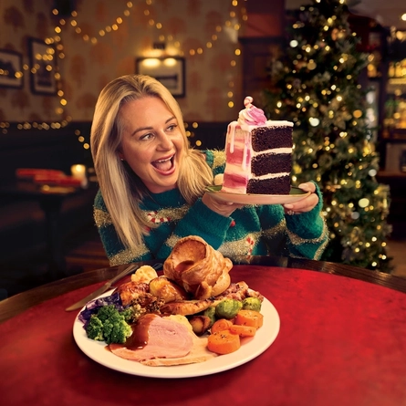 A roast dinner featuring roast meat, Yorkshire pudding, vegetables and gravy, served on a plate which sits on a table. A woman is sitting at the table holding a large slice of cake on a plate.