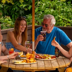 A mixed group of people seated around a wooden table, under an umbrella in a beer garden, enjoying a variety of sharers and drinks.