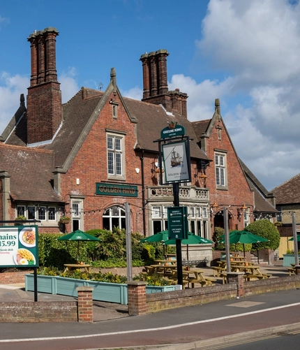The exterior facade, signage, and beer garden seating area of the Golden Hind in Cambridge, with wooden picnic tables, shade umbrellas, and string lights.