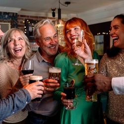 Six people gathered closely together indoors, each holding a small shot glass during a celebratory toast. One person wears a green outfit and another a silver sequined top. The group stands in a warmly lit venue, with drinks raised and decorative lighting visible in the background.