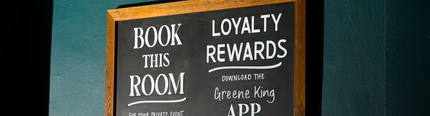 A chalkboard sign on a dark teal wall inside the Coach & Horses, promoting the Greene King App, and a room available to book for private events.
