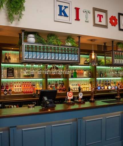 The bar inside The Kittoch in East Kilbride, with potted plants and bottles of wine on shelves above the counter, and bottles of alcoholic spirits on illuminated shelves behind the bar.