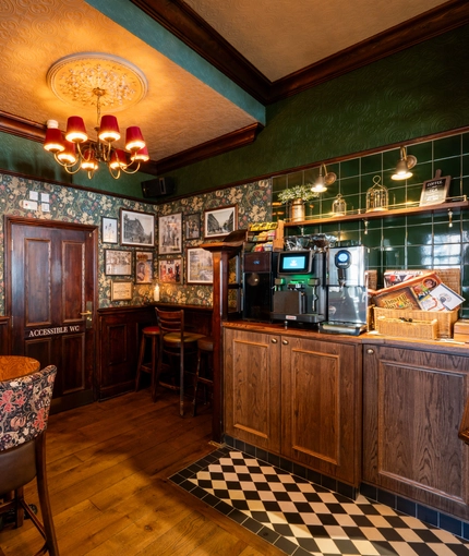 An interior restaurant seating area at the King's Head, with dark wood floors; floral patterned wallpaper and green tiles on the walls; and a coffee machine.