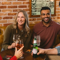 People gathered around a pub table with a glass of white wine, a bottled beer and other drinks, against a warm exposed-brick backdrop.
