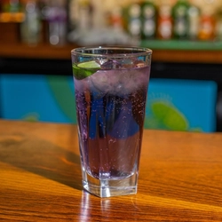 A close up view of a JD Blackberry & Tropical Soda cocktail, served in a highball glass with ice and a wedge of lime. The glass stands on the bar inside a pub.