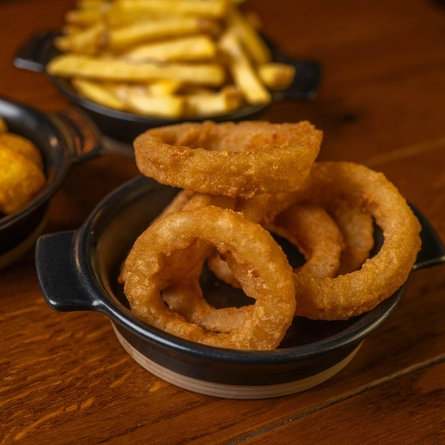 A close up view of Onion Rings served in a small roasting dish which sits on a wooden restaurant table along with dishes of fries and tater tots.