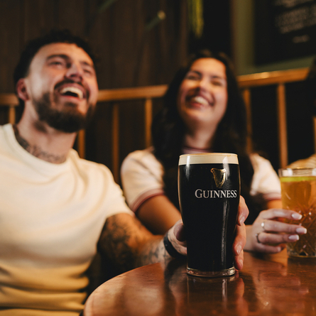 A pint of Guinness on the table in the interior seating area of a Crafted venue, three people are blurred in the background, one is holding the pint.