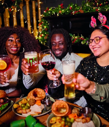 A group of people celebrating a festive Cheers while enjoying various main meals and drinks within the interior restaurant and seating area of Greene King Pub venue.