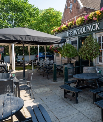The exterior facade, signage, and beer garden seating area of The Woolpack Inn; with wooden picnic tables, trees on each side of the entrance, and a large shade umbrella.