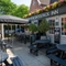 The exterior facade, signage, and beer garden seating area of The Woolpack Inn; with wooden picnic tables, trees on each side of the entrance, and a large shade umbrella.