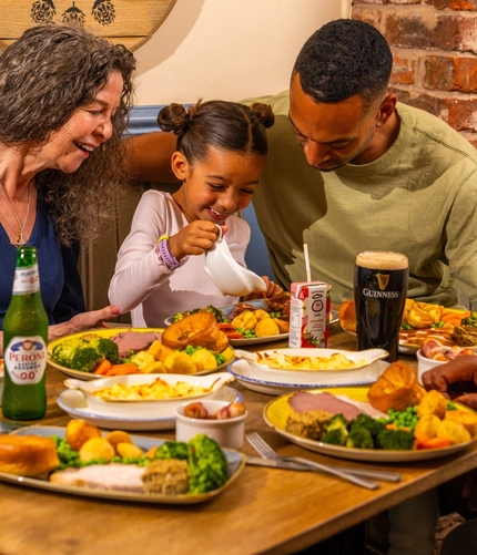 Three people seated around a wooden table in a pub, sharing Sunday roast meals. One person pours gravy from a small jug onto a plate of roast dinner with Yorkshire puddings, vegetables and potatoes. A bottle of alcohol-free beer, a pint of Guinness and a children's juice carton sit among the dishes. The background features blue panelled walls, exposed brickwork and a wooden barrel-style sign reading “The Mill House”.