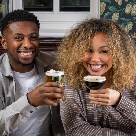 Two people sit together in a restaurant booth. They are raising their drinks toward each other, one holding a pint of Level Head, the other holding an Espresso Martini in a stemmed glass. A wooden table in front of them is set with shared dishes, including loaded nachos topped with cheese and colorful toppings, and a plate of fried appetizers with dipping sauce.