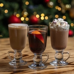 Three festive hot drinks served in Irish coffee glasses. The glasses sit on a wooden table and a Christmas tree is visible in the background.