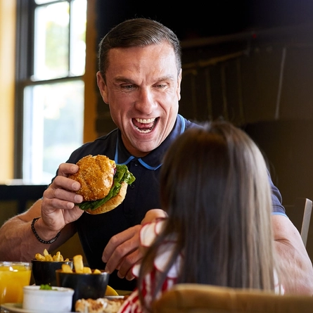 A man seated at an indoor table, talking to a child and enjoying a burger.