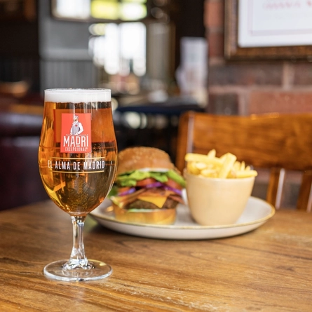 A served pint of Madri in the interior restaurant and seating area of a Community Pubs with Food venue, a blurred burger meal sits in the background.