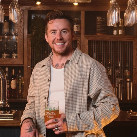 Danny Jones stood next to a bar, smiling at the camera whilst holding a pint of Level Head Ale.